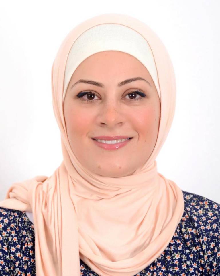 Image of a woman staring at the camera, smiling, wearing a light peach headscarf over a white undersarv and a dark blouse.