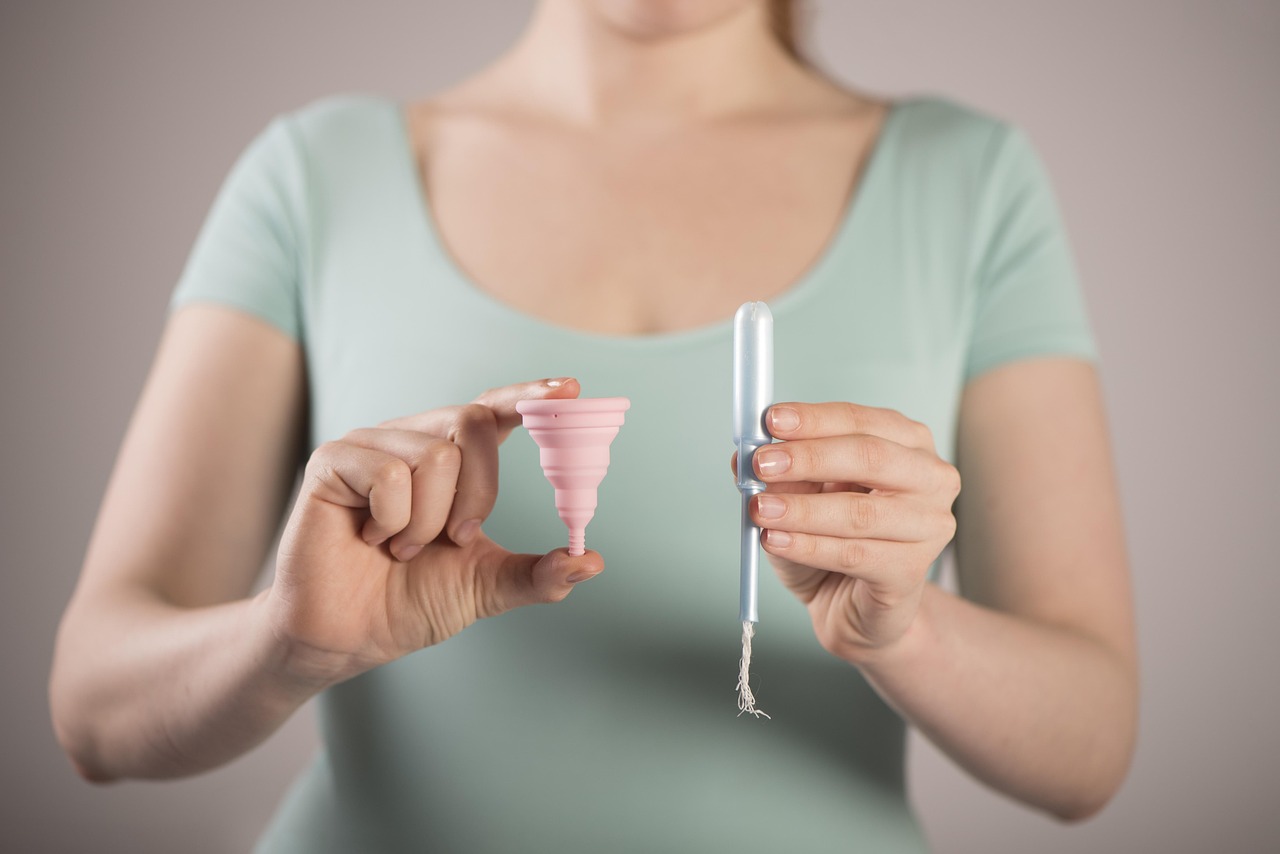 Image of a woman from shoulders to mid abdomen. Her hands are in front. Her right hand is holding a reusable pink menstrual cup. Her left hand is holding a disposable tampon. She is wearing a light green t-shirt.